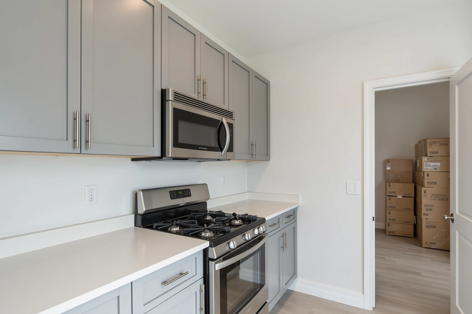 Grey shaker apartment kitchen with a white quartz counter and stainless range, cardboard boxes stacked beyond the doorway.