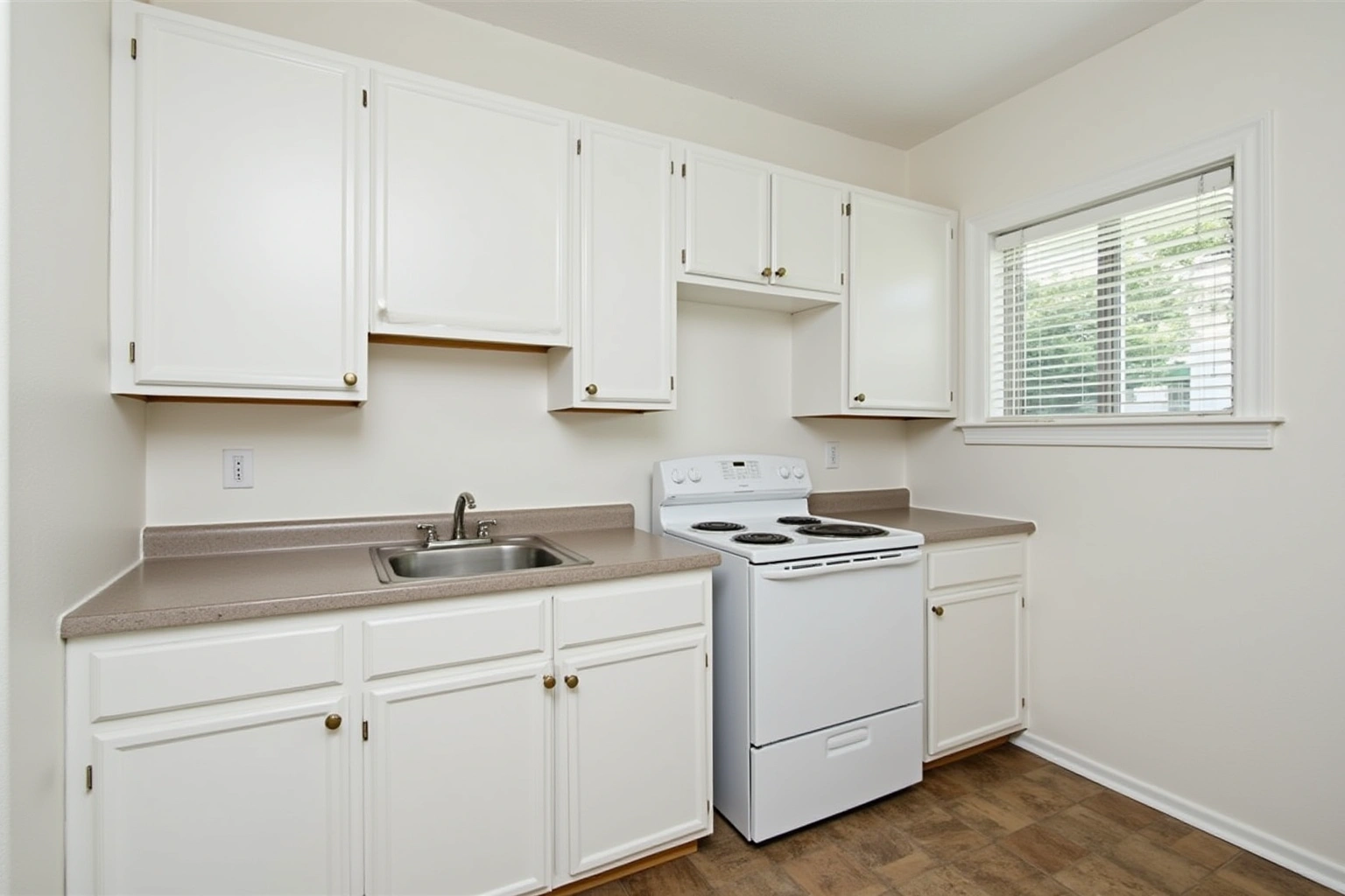 Simple white raised-panel stock cabinets with a tan laminate counter, white range and a window with blinds.